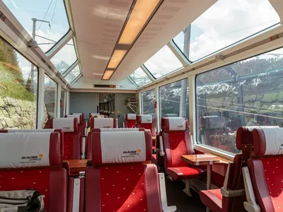Inside a Glacier Express first-class panorama car in 2014, with red seats in pairs either side of small tables and windows curving up into the roof.