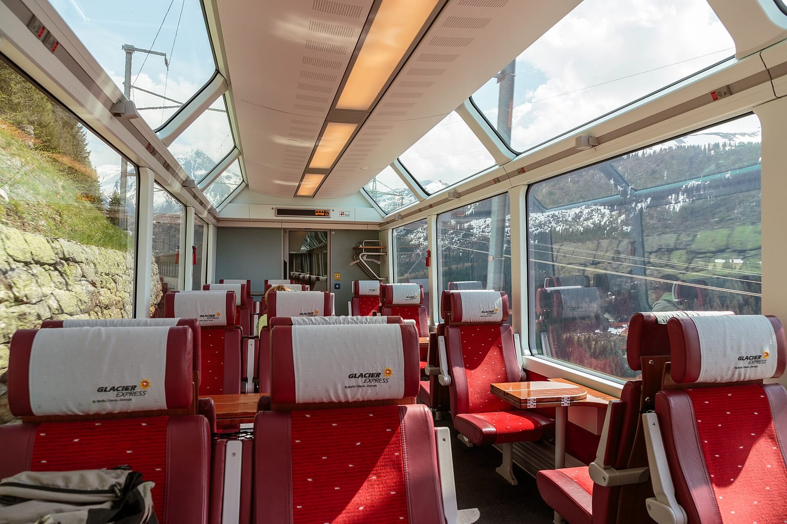 Inside a Glacier Express first-class panorama car in 2014, with red seats in pairs either side of small tables and windows curving up into the roof.
