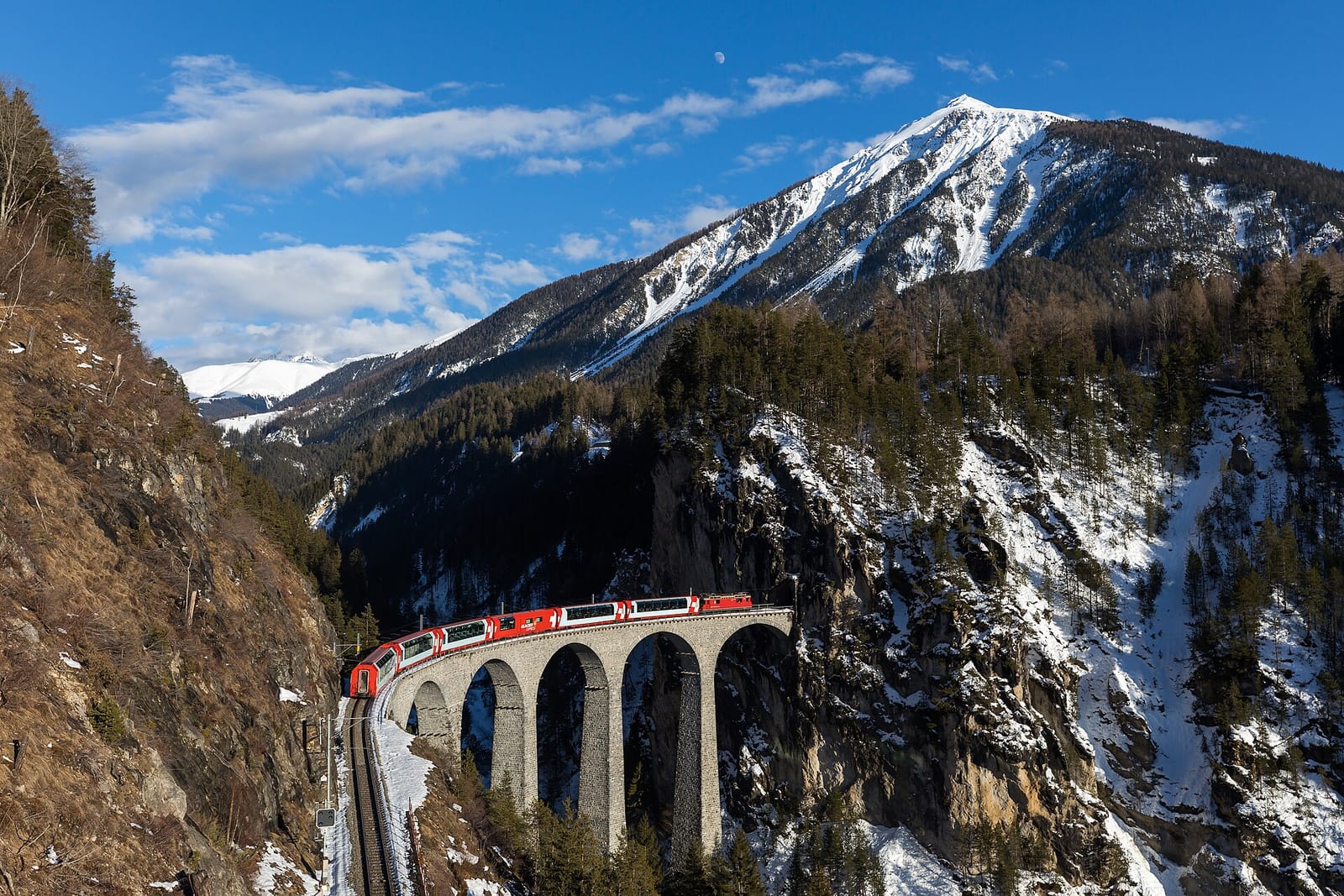 A red Glacier Express train, hauled by a Rhaetian Railway locomotive, crossing the Landwasser Viaduct below snow-covered mountains in winter.