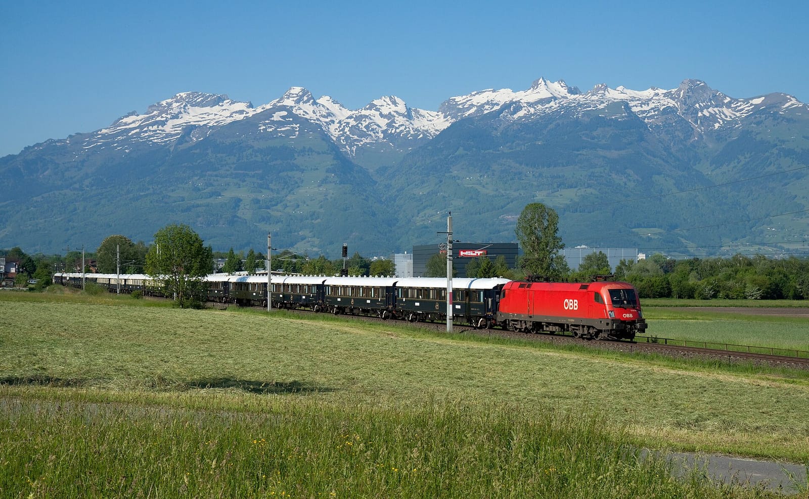 The Venice Simplon-Orient-Express, hauled by a red ÖBB locomotive, passing fields at Nendeln in Liechtenstein, with snow-capped mountains behind.
